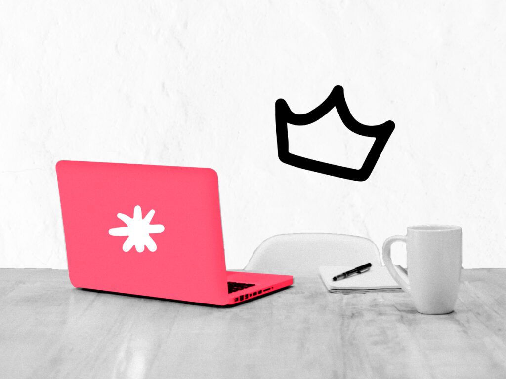 A laptop on a desk with a mug and a crown over the chair